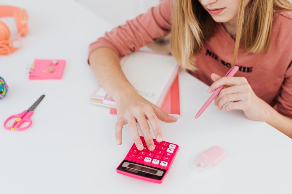 Teenage girl using a pink calculator while studying at a desk with school supplies.