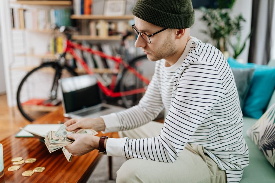 Man in striped shirt counting cash indoors, with a bicycle in the background.