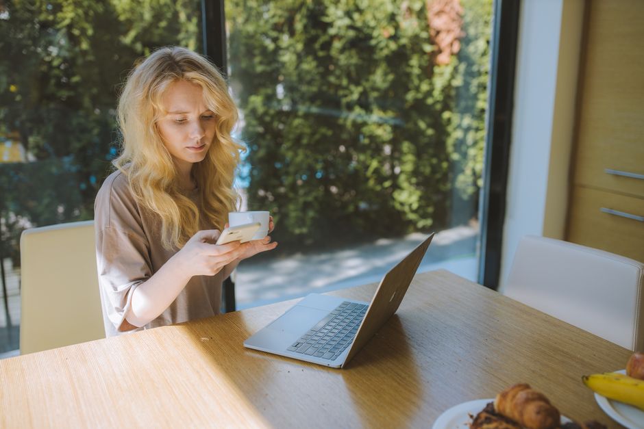 Blonde woman using smartphone while working on a laptop at home by a large window.