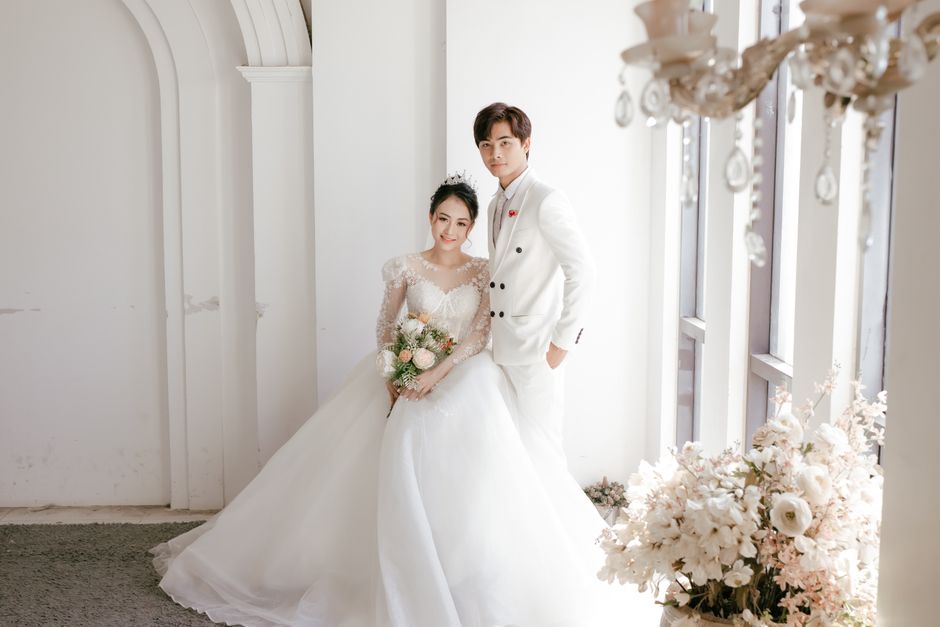 Full body cheerful newlywed Asian couple wearing wedding gowns standing near window in light luxury mansion and looking at camera